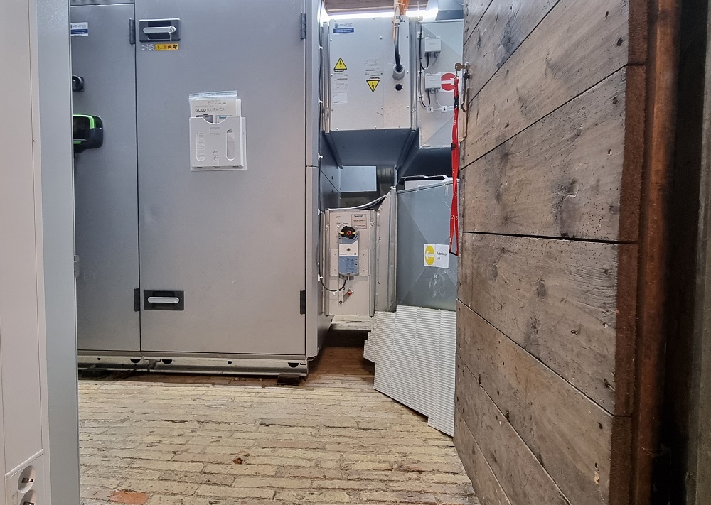 Det grå ventilasjonsaggregatet står på det originale, lyse mursteinsgulvet i det lille huset som utgjør kjernen av første etasje. Foto.