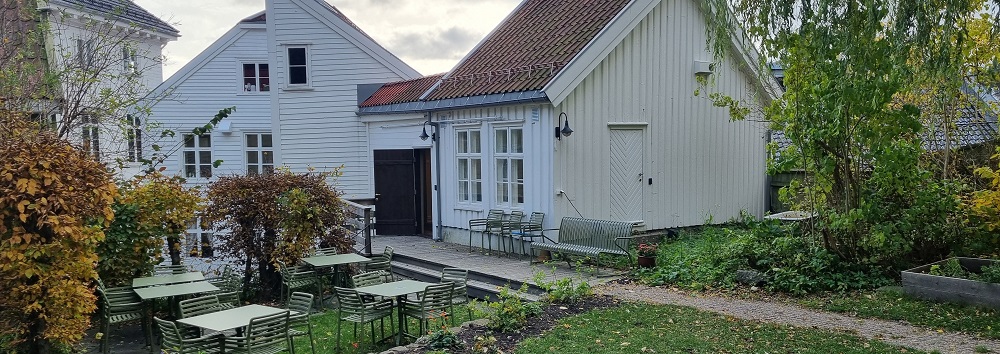 Bakgården med grønt gress, gangsti fra døra, fire benker med bord, stoler, urtekasser og prydbusker. Foto.