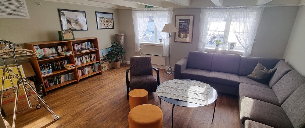 Dagligstue i andre etasje med grå hjørnesofa, bokhylle, et rundt bord, en brun stol og to orange krakker. To vinduer med blondegardiner og sjøutsikt. Foto.