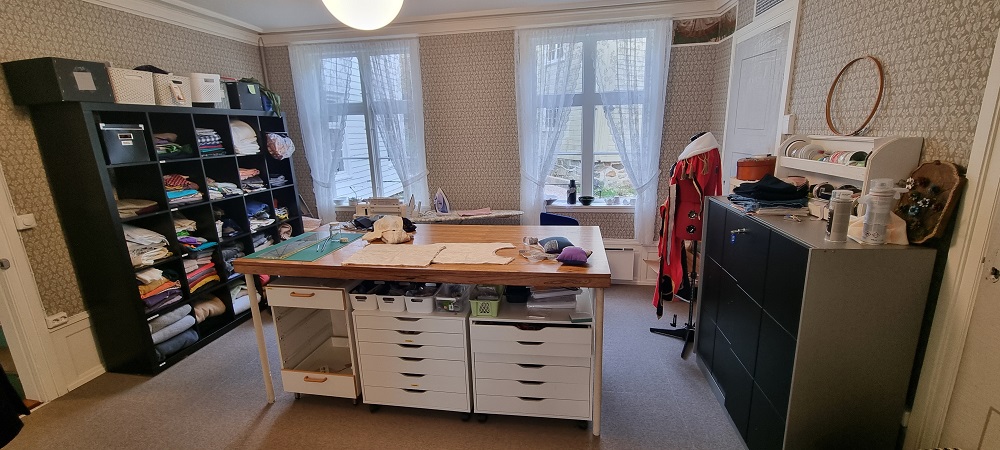 Systue rom, sorte reoler på to sider med stoff, stor øy midt på gulvet med skuffer, mønster og skjæreutstyr. To stoe vinduer og høyt under taket, Foto.