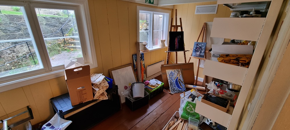 Atelier, avlangt gult rom, hvite hyller med malerutstyr, staffili, lerret og rammer på gulvet. To vinduer ut mot bakgården, brunt gulv. Foto.