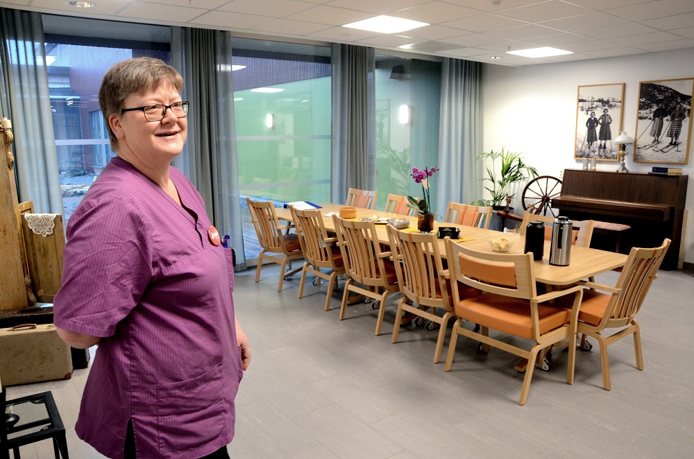 Enhetsleder avbildet i fellesrom med stoler, bord, gamle bilder, en rokk og et piano. Foto. 