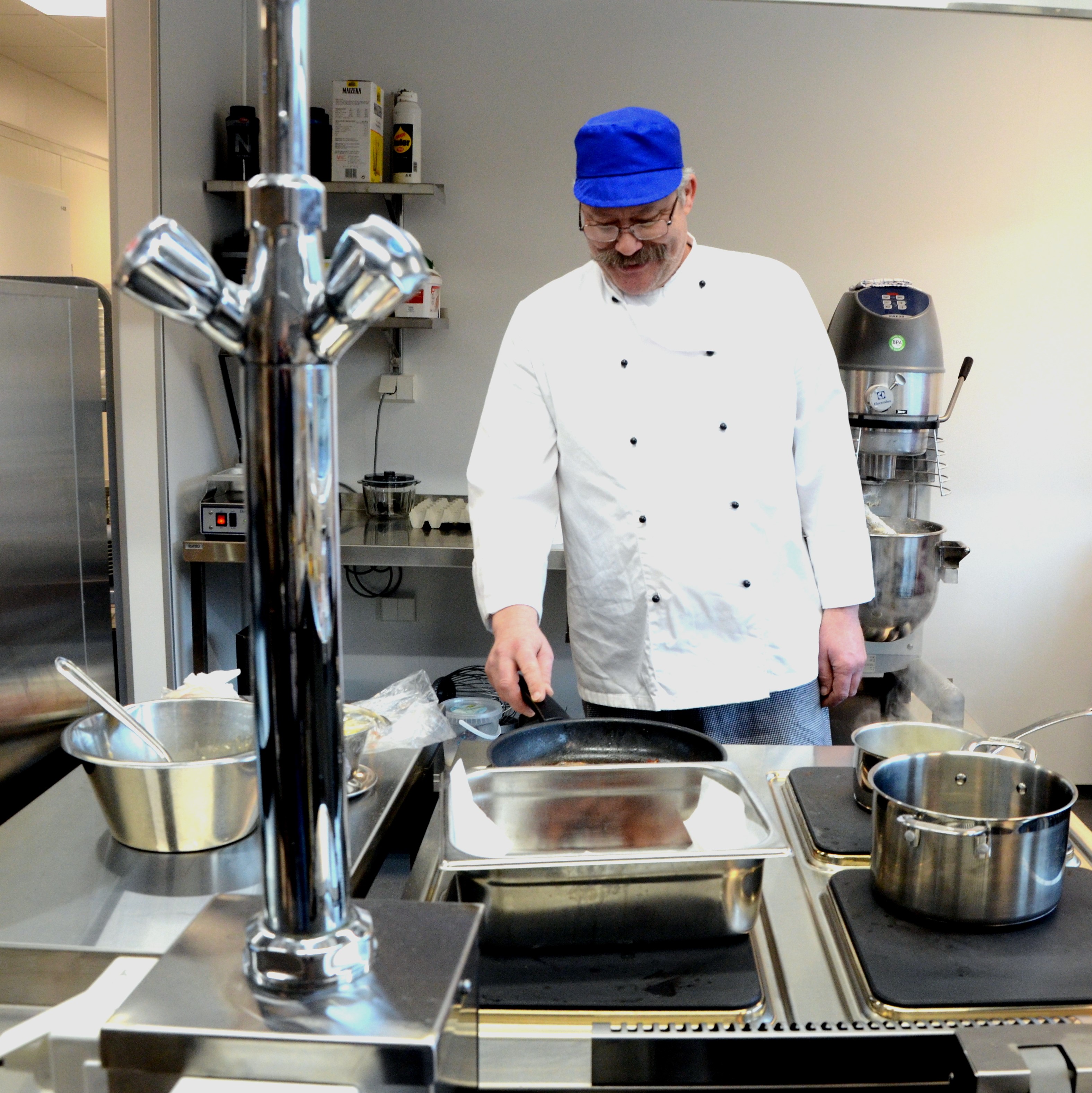 Kokken med hvit jakke og blå lue avbildet på kjøkkenet. Foto. 