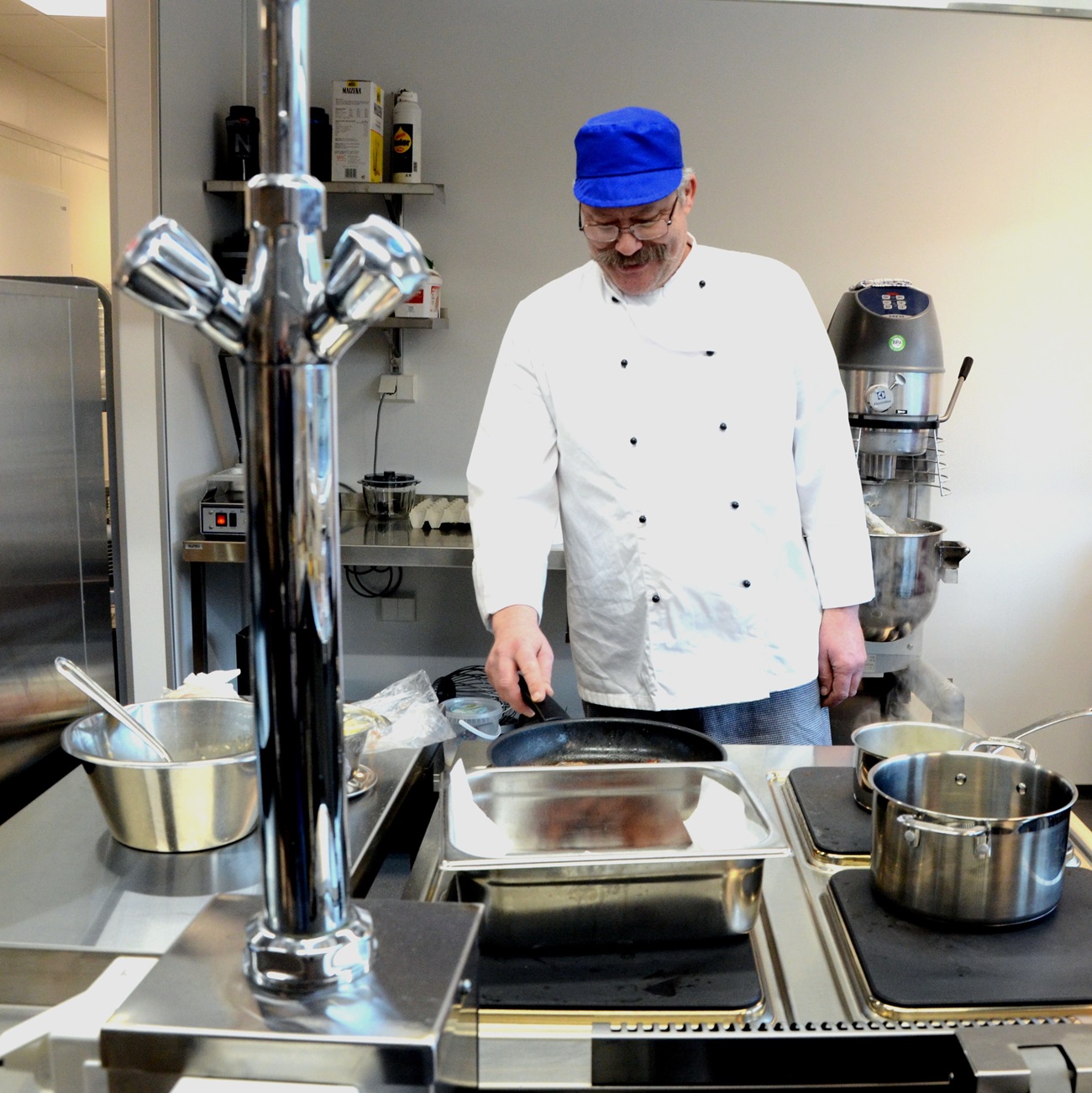 Kokken med hvit jakke og blå lue avbildet på kjøkkenet. Foto.