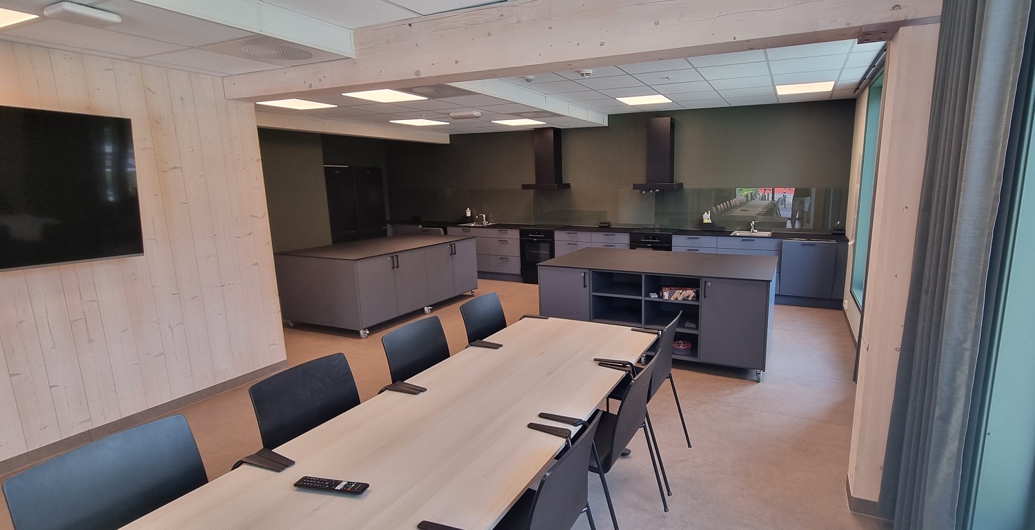 Stort fellesrom til utlån i det ene bygget med langbord og stort, koksgrått kjøkken. Mørke grønne kontrastvegger på kjøkkenet og gardiner, ellers lyse massivtre vegger. Foto. 