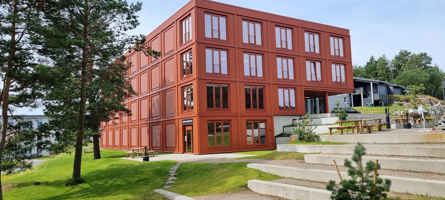 Rødt hybelbygg sett fra tunet. Grønt gress, amfitrapper, sittebenker og noen trær til venstre. Foto. 