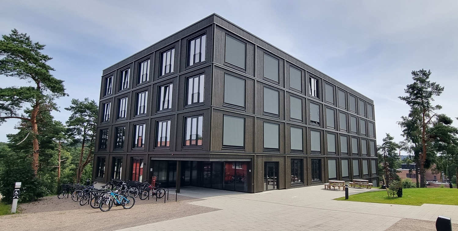 Brunt hybelbygg sett mot inngangsparti. Hellelagt område med sykkelparkering og litt grønt gress og trær på sidene. 