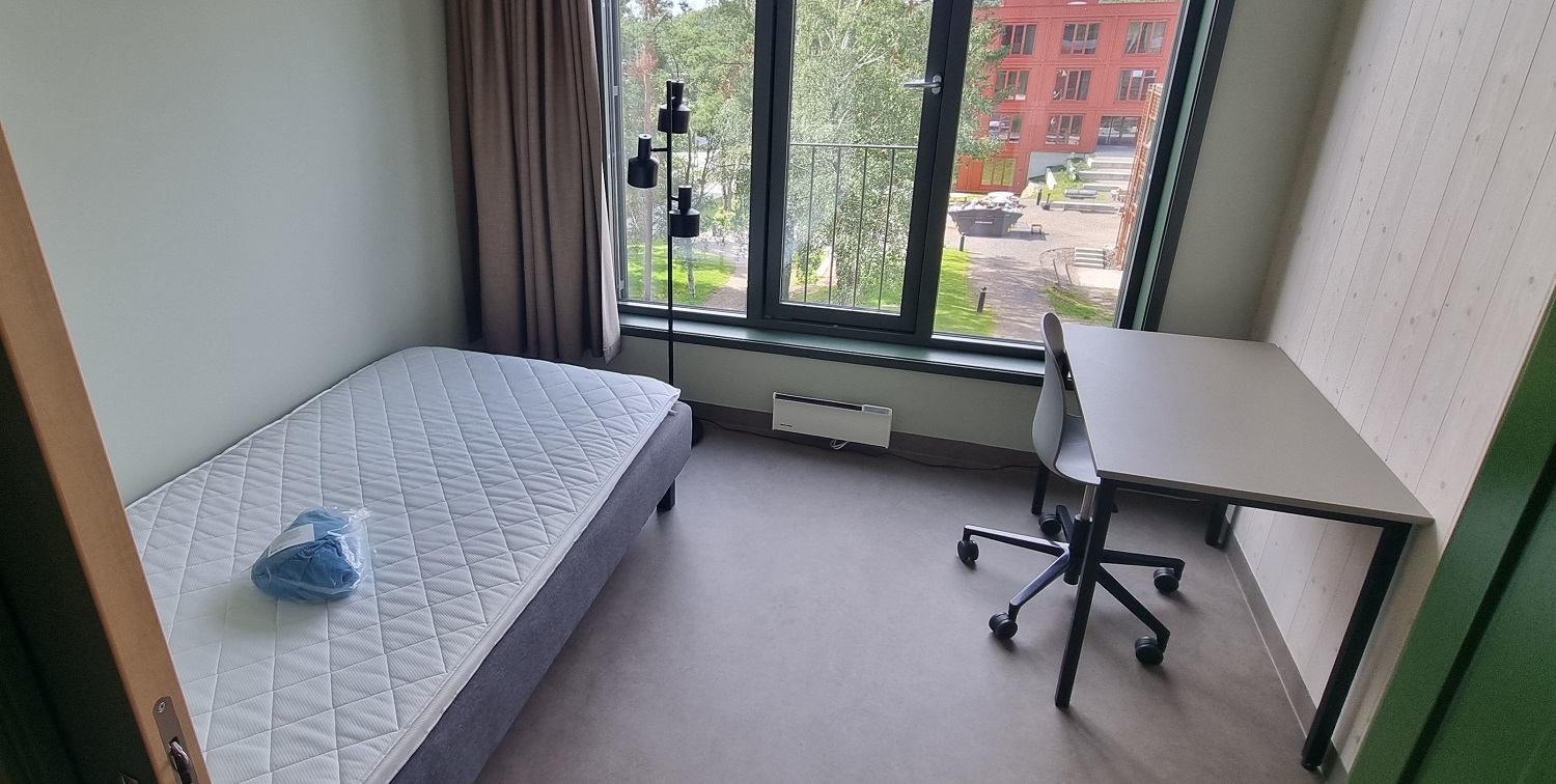 Rom med hvite vegger, seng, høye vinduer med utsikt over tunet og en pult med kontorstol. Foto. 