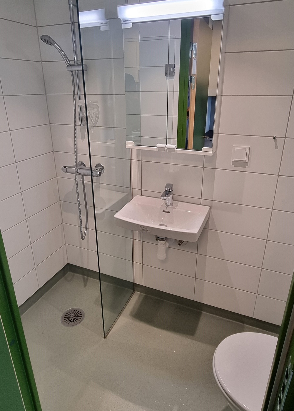 Hvitt bad med veggfliser og gulvbelegg. Dusjkrok med glassvegg, hvit servant med speil og gulvklosett. Foto. 