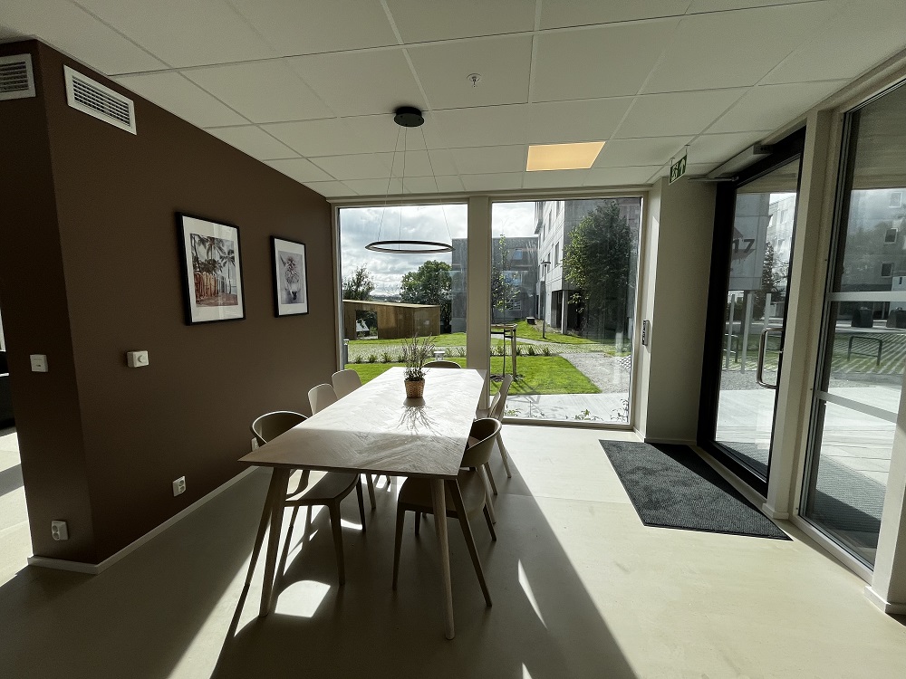 Innvendig fellesområde på bakkeplan med bord, stolar og store vindaugsflater. Foto. 