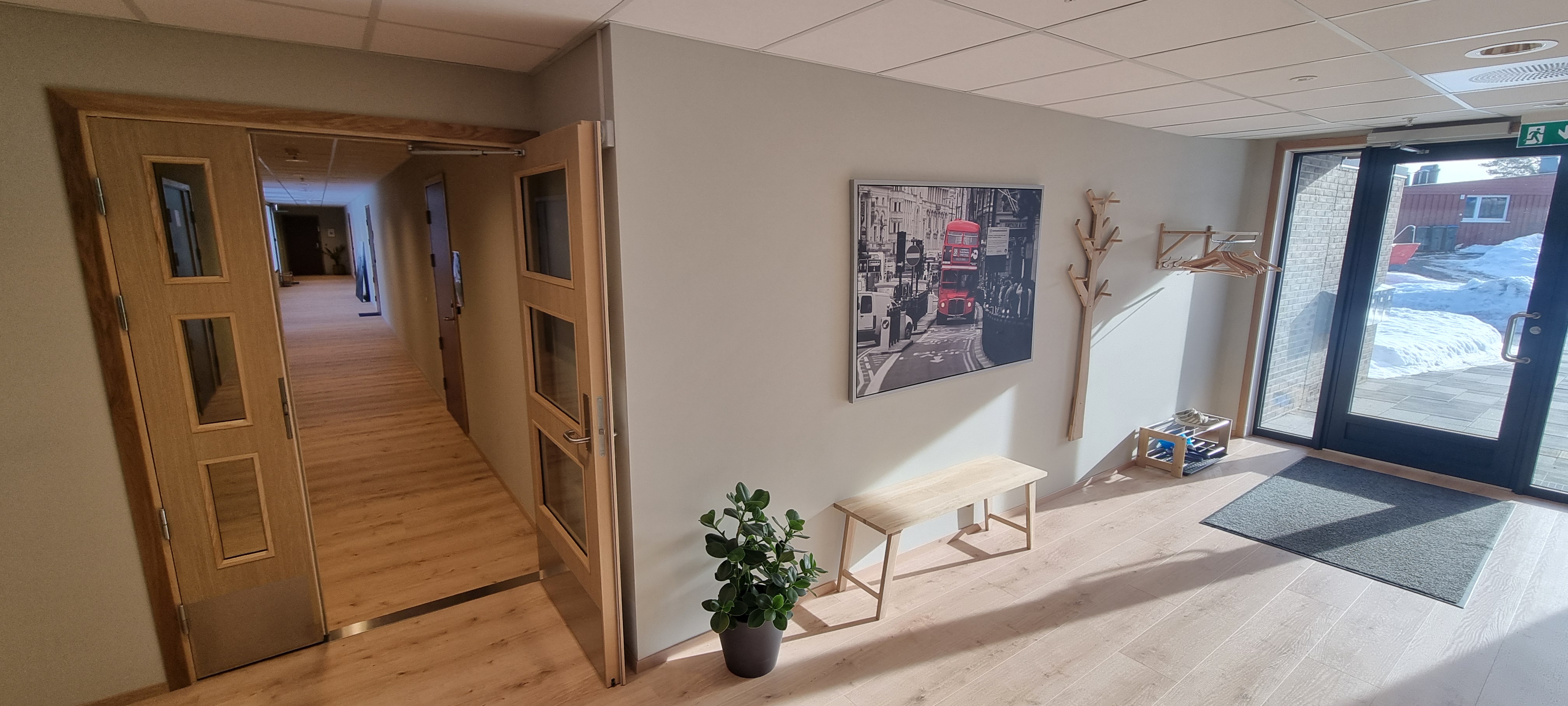 Stor, felles entre med lyse vegger og parkett, fargerikt maleri, skohylle og kleshenger. Åpen dør med sidefelt inn til svalgang og de fleste leilighetene. Foto. 