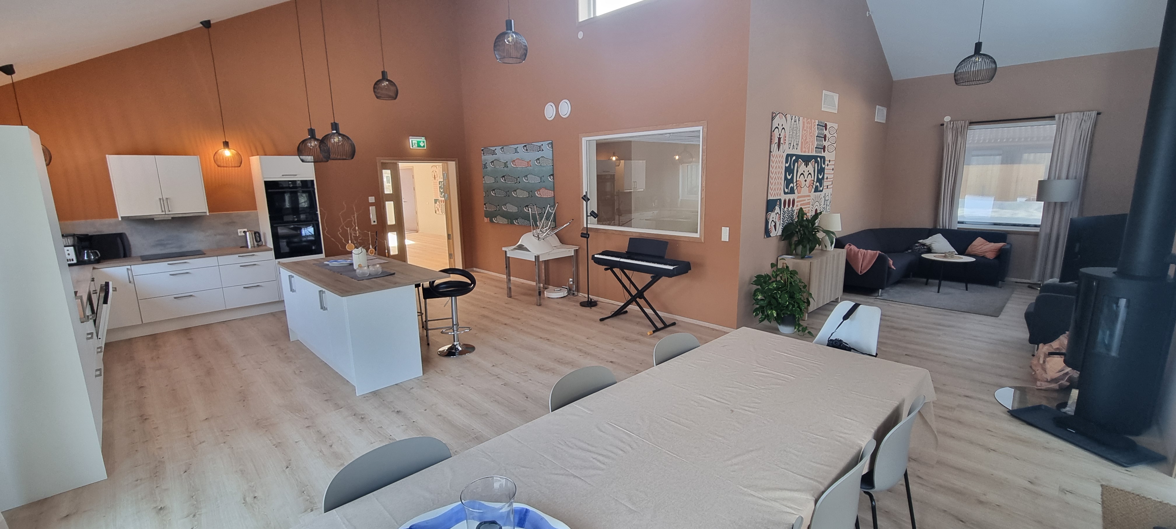 Stort fellesrom med varme farger og god takhøyde. Sofakrok med TV, peis, Stor kjøkkenkrok med hvit innredning og øy. Spisebord med 12 stoler. Keyboard under vindu inn til treningsrom. Foto.