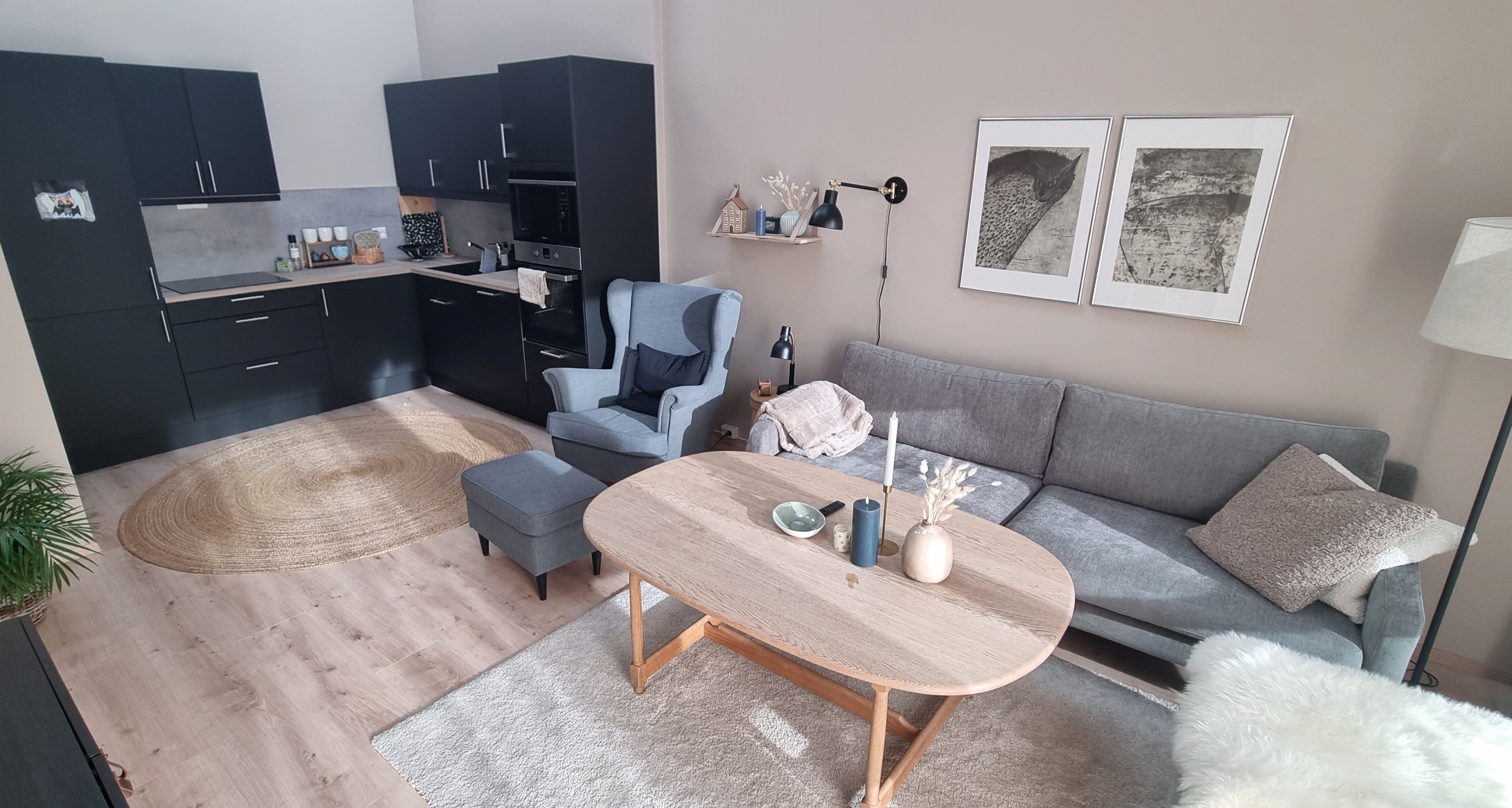 Åpen løsning med mørkt kjøkken og stue med lyseblå sofa og ørelappstol, stålampe og stuebord. Lyse og varme farger, To bilder på veggen over sofaen. TV. Foto. 