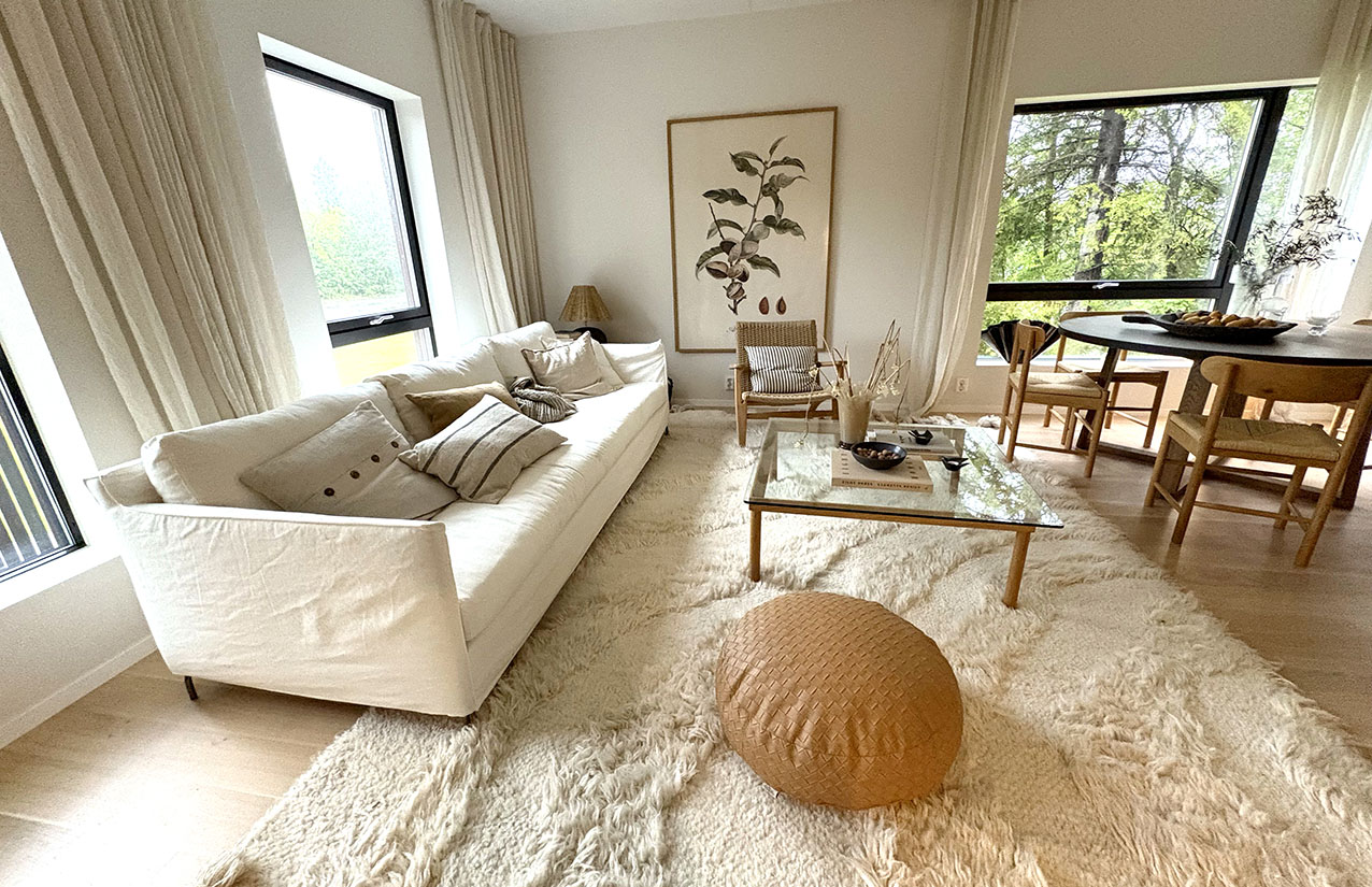 Stue med lys sofa, glassbord og spiseplass ved store vinduer.