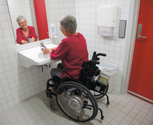 Dame i rullestol som vasker hendene og ser seg i speil over servanten. Foto.
